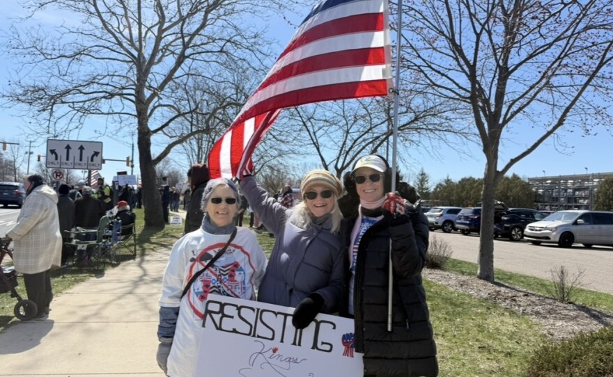Three women hold a sign that says "resisting Kings since 1776, signed we the people". One holds a large American flag. 
