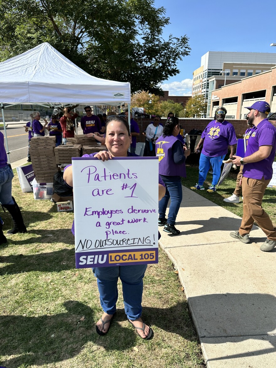 Deanna Gonzales stands holding a white sign that reads "Patients are #1" with other strikers wearing purple shirts walking in the background. 