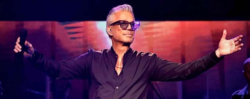 Man with grey hair on stage wearing black shirt and sunglasses holding a microphone with his arms open 