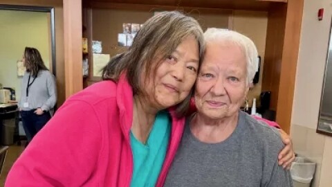 Two women stand close together, one holding a plate with bread and a bowl of soup.