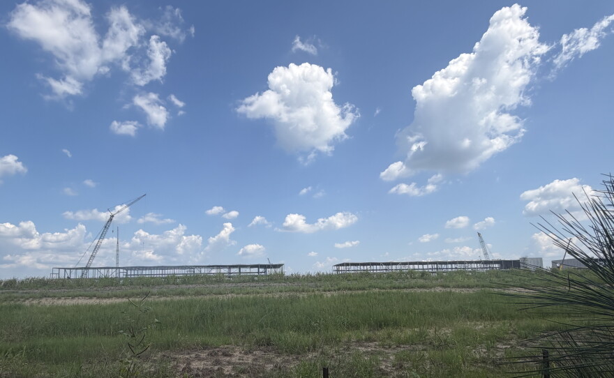 There is grass in the lower third of the photo. In the middle are two steel structures - the skeletons of what will become buildings for the data center. The upper part of the photo is a clear blue sky with some clouds.