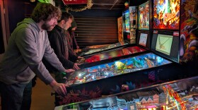 Three men stand at pinball machines playing pinball,