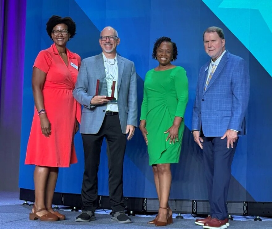Four people stand onstage posing for a photo, with one man holding an award.