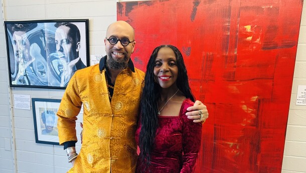 A man in a yellow jacket and woman in a black velvet dress stand in front of a big red canvas shoulder-to-shoulder. 