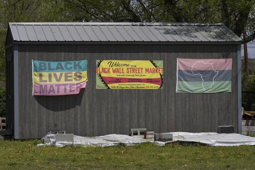 A dark-colored shed has three flags hanging on it -- one reads "Black Lives Matter" and another welcomes visitors to the Black Wall Street Market, and the other has an outline of Africa against red, black and green horizontal stripes.