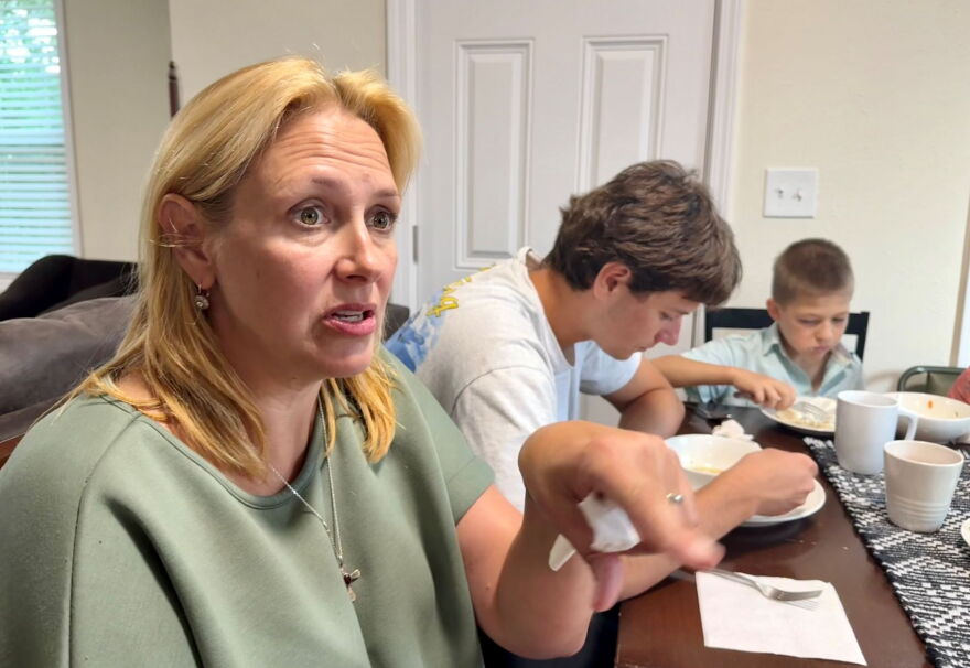 A blonde woman in a mint-green shirt chats at a dinner table. Two kids are visible in the background. 