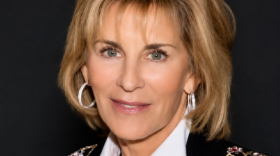 A smiling woman with short blonde hair, wearing white hoop earrings, a necklace, and a white blouse under a black blazer with hearts and stars embroidered on it.