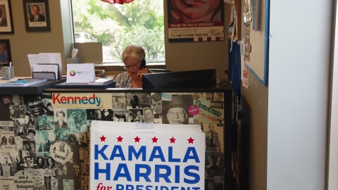 A volunteer with the Kalamazoo County Democratic Party sits at a desk covered with different pictures of past democratic candidates and presidents. She's on the phone, with other political posters other democratic advertisements behind her on the wall. In front of the desk is a white sign promoting Kalama Harris' presidential campaign. At the top of the sign is six red stars, the sign reads "Kamala Harris for President."