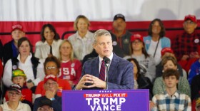 U.S. Representative Bill Huizenga at a campaign rally is at a podium with a Trump Vance campaign sign on the front of it.  Behind him is a crowd of supporters.  Huizenga wears a blue plaid sports coat, khaki pants, blue and red stiped tie and a white dress shirt.