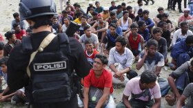 A police officer stands guard as ethnic Rohingya men sit on a beach after they landed in Aceh Besar, Indonesia, Sunday, Jan. 8, 2023. The group of more than 100 hungry Rohingya Muslim refugees were among the latest group to reach ashore in Indonesia on Sunday after a long and dangerous journey aboard a wooden boat.
