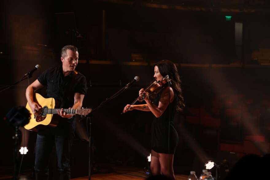 A man playing an acoustic guitar and a woman playing a violin perform on a darkened stage.