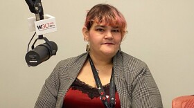 A person with short, wavy, brown and pink hair sits next to a microphone labeled "WGLT." They wear a plaid blazer, a black and burgundy top, earrings, and a lanyard, seated in a studio setting.