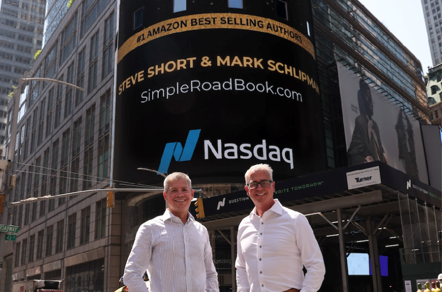 Two men in button up shirts pose for a photo in front of an advertisement display for Amazon best selling authors Steve Short and Mark Schlipman 