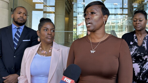 A photo of Crystal Mason, a Black woman with short, straight black hair, wearing a brown dress and a butterfly necklace. She speaks into a microphone at a press conference, surrounded by her attorneys and supporters.
