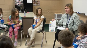 A group of children sit in a circle in a classroom, looking at a paper about feelings held up by a woman