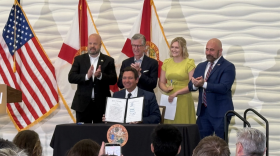 Un hombre de traje sonríe y muestra un documento a una multitud. Tres hombres de traje y una mujer con un vestido amarillo están de pie detrás de él.