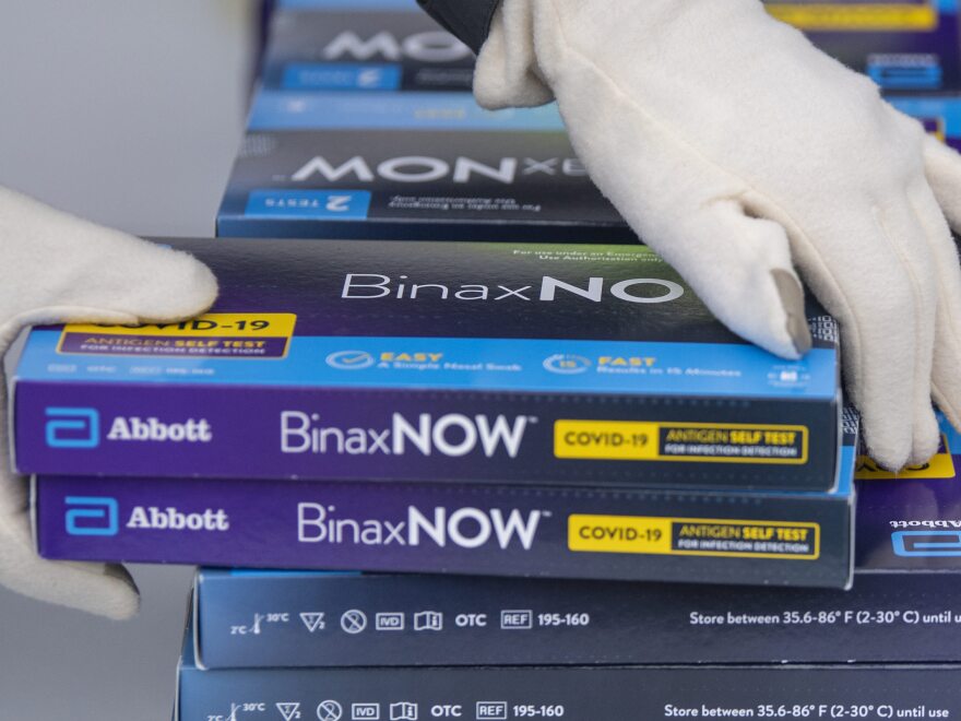 Gloved hands grasp blue boxes of BinaxNOW COVID-19 tests