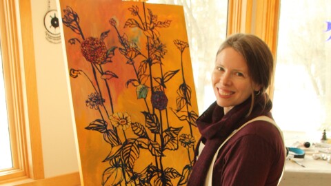 A woman wearing maroon in front of a yellow painting of flowers.