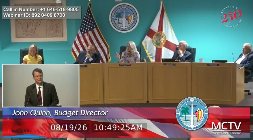 John Quinn appears in a tiny window on the lower-left of the screen with Monroe County Commissioners appearing on the dais behind him.