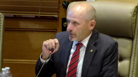 A man in a suit and red striped tie sits at a conference table, speaking into a microphone. He appears to be in a formal meeting room with a wooden door and leather chair visible in the background.