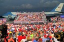 huge crowd of people primarily wearing red while Trump is in the center talking to them. Very large plane in the background, starting to turn into nighttime