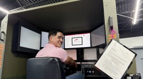 A young man in a pink polo sits inside a airplane flight simulator. He has his head turned looking back and is smiling. In front of him is a digital view of a runway. 