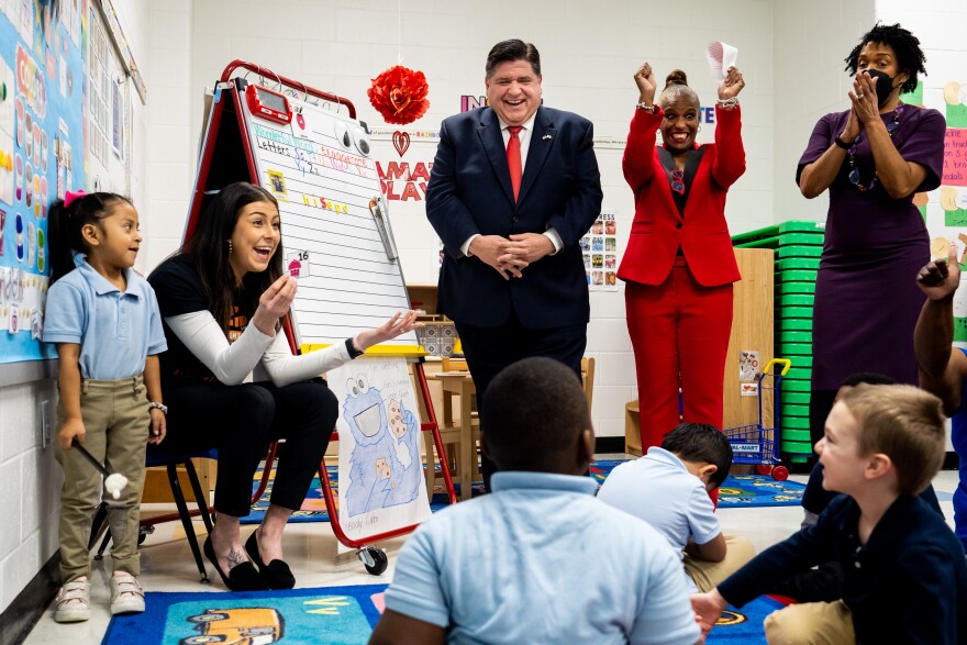 A teacher goes over counting with her students sitting on a rug as Gov. J.B. Pritzker, Principal Melanie Hood, and Lt. Gov. Juliana Stratton cheer