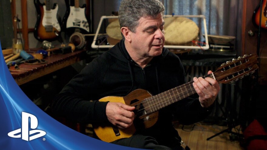 composer Gustavo Santaolalla sits in studio surrounded by musical instruments, holding a small guitar-like 10-stringed instrument