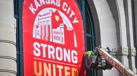 Two men can be seen high off the ground in a bucket lift that is extended toward the outside of a large, concrete building. They appear to be hanging a large red banner with white and gold letters that reads "Kansas City, Strong, United." 