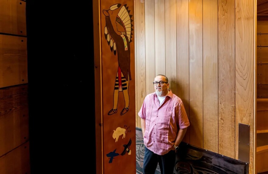 A man stands against a wooden wall and looks at the camera. One panel has a painting of a Native person in regalia. 