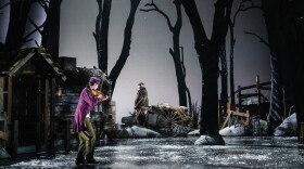 A young man wearing a purple coat and cap plays the violin on stage. In the background, an older man, wearing a villager costume, gazes at the violonist, while pulling a cart.