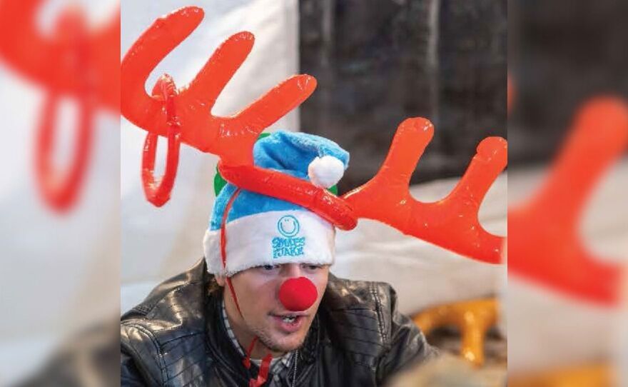 A man with large red reindeer antlers and a sky blue Santa hat and a clown nose looks ahead.