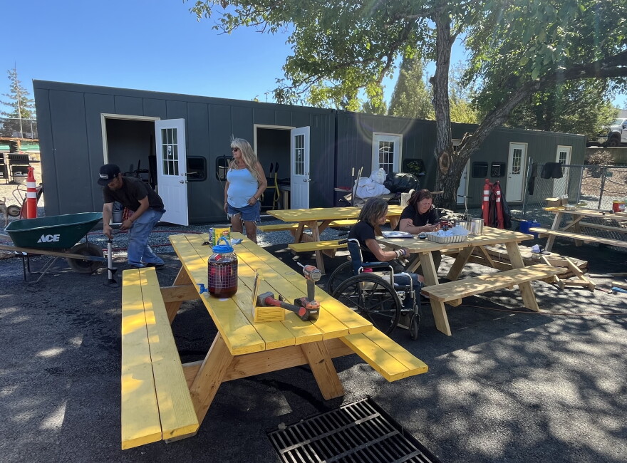Picnic tables are shown on pavement under the shade of a large tree. A few people are seated at the tables. 