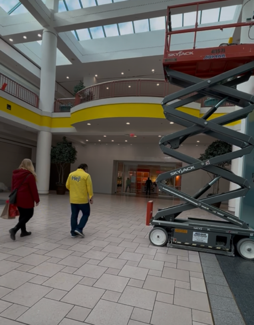 A construction lift inside a mall at the entrance of a new IKEA store, with two people walking toward the store. 