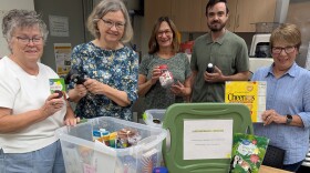 Four older white women and one younger white man stand around a plastic bin full of shelf-stable food, like Cheerios and canned goods. They're smiling and posing with the items.