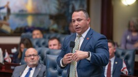 Man in suit and tie speaks into microphone with other representatives around him 
