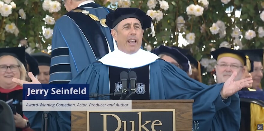 Jerry Seinfeld, wearing a graduation cap and gown, gesturing behind a podium that reads 'Duke'