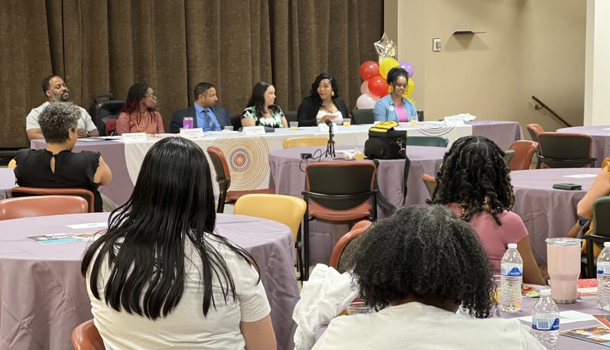 A large conference room has several round tables with purple table clothes and women sitting at them. A group of speakers sit at the front of the room at a rectangular table with red and yellow balloons and a brown curtain behind them.