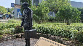 A statue of a young man with a suitcase and cane is visible in a park in downtown Petoskey. In the foreground, a gold plaque reads "Ernest Miller Hemingway." 