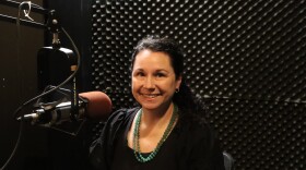 A woman sits smiling in a recording studio beside a microphone.