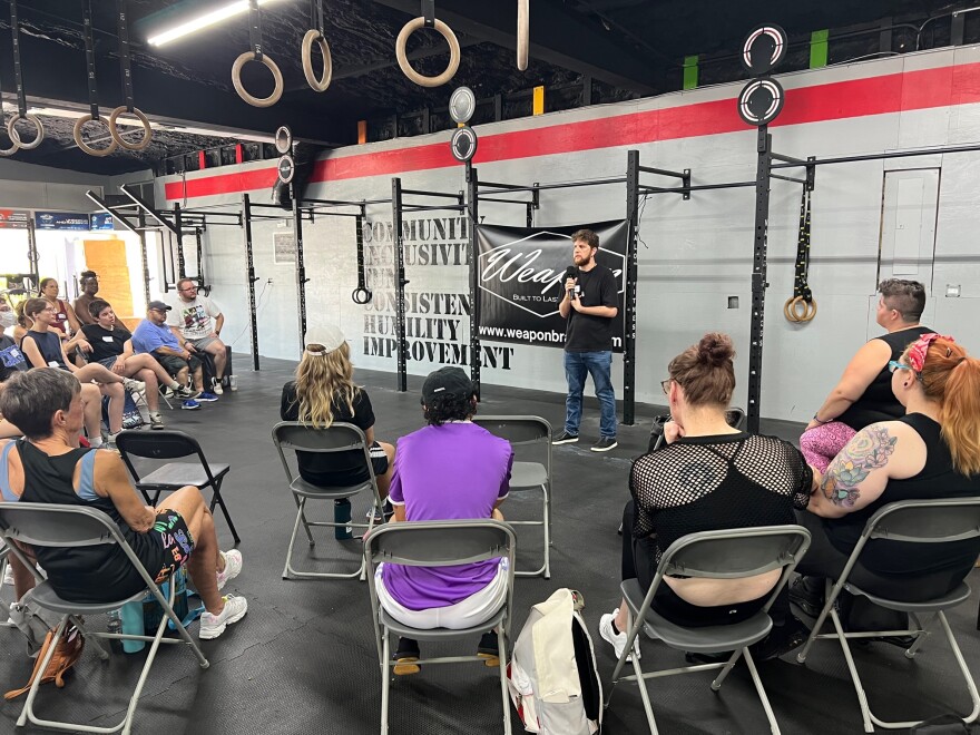 Group of people sit in folding chairs in a gym. Man stands in front of them and addresses them with a microphone.