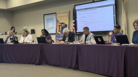 District 87 school board members and the superintendent sit at a long table draped in purple with laptops and nameplates, beneath a screen showing the Oct. 21 meeting agenda and beside a Bloomington Public Schools banner.