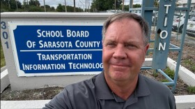 A gray haired man in gray collared shirt takes a selfie by a sign that reads "school board of Sarasota County."