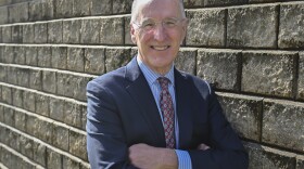 A man wearing a blue suit and red tie leans on a brick wall with his arms folded. He is smiling and looking at the camera.