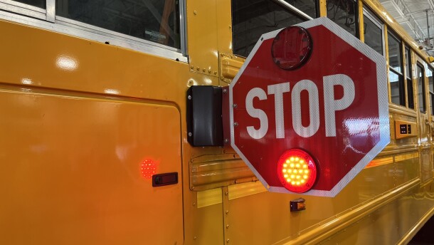 The side of a yellow school bus with a red stop sign extended.  Visible behind the stop sign is a housing for cameras that are intended to catch drivers illegally passing the bus.