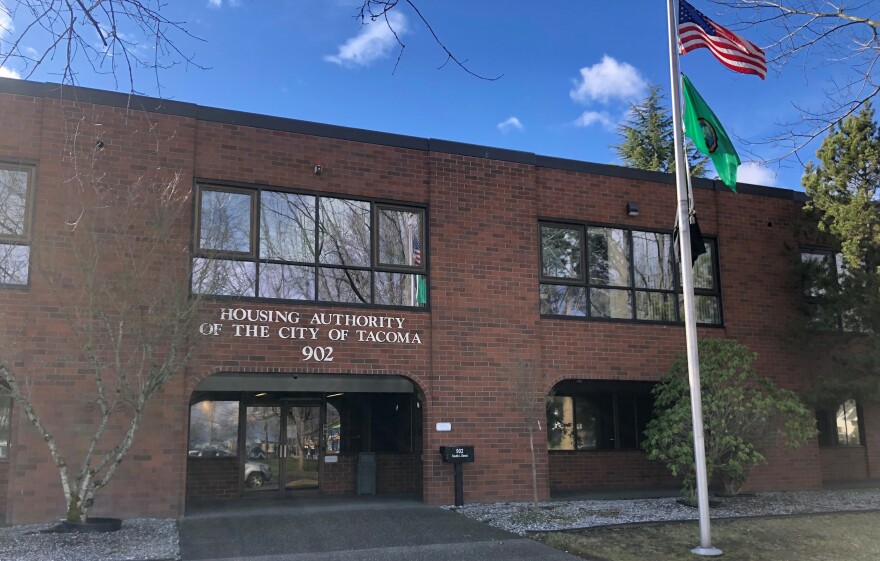 The outside of a brick building with a sign that reads "HOUSING AUTHORITY OF THE CITY OF TACOMA 902". There's flag staff to the right of the entrance of the building with an American flag and Washington State flag.