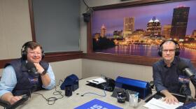 Two men wearing headphones and eyeglasses sit at a table in a radio talk studio: a man at left has short brown hair and is wearing a navy blue puffy vest over a light blue button-down shirt; a man at right has very short dark hair and is wearing a navy button-down shirt.