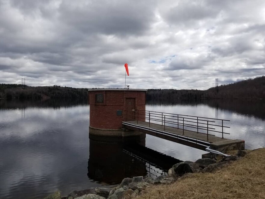 A small round brick building with a little orange flag on its roof sits in a body of water under swollen grey clouds