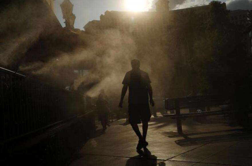 A person, in silhouette, stands in a plaza as misters activate around them. The sun can be seen, big, bright, and shining above the person.
