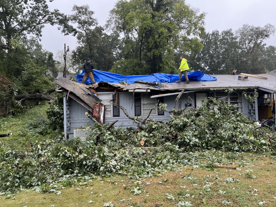 Two men on a house's roof stretch a blue tarp across an opening.